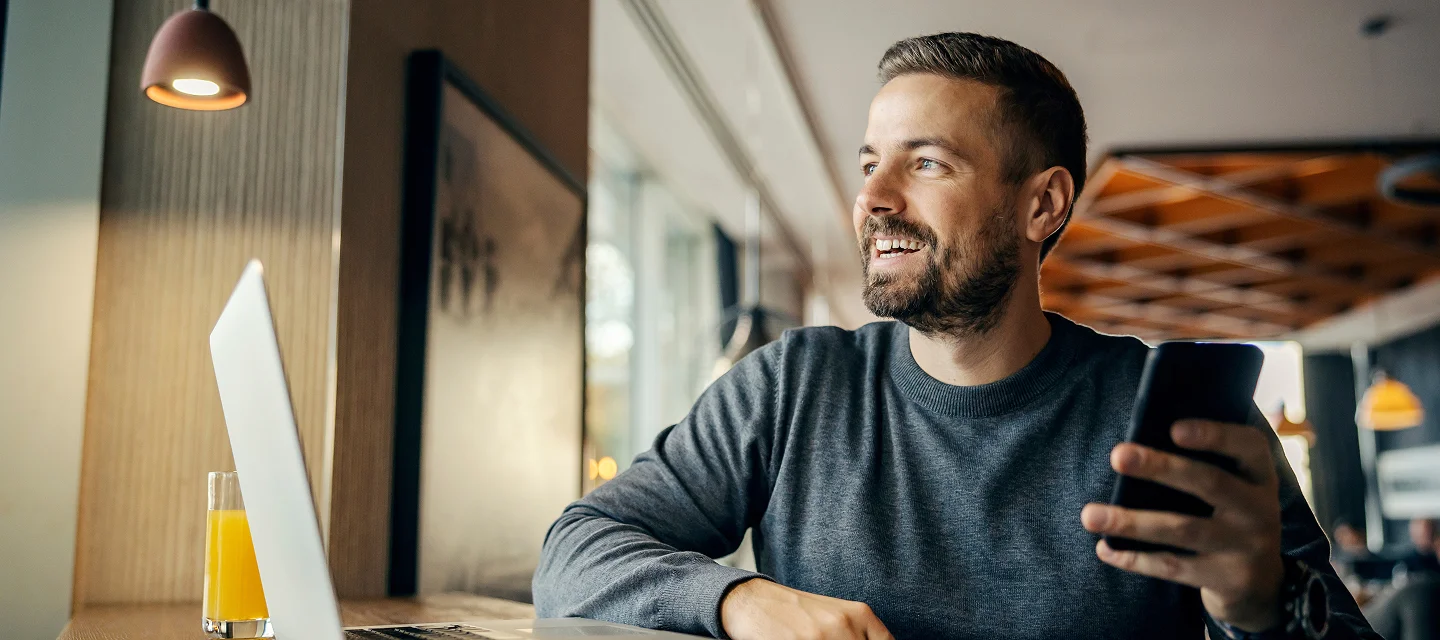 Ein lächelnder Mann sitzt mit einem Smartphone in der Hand an einem Tisch vor seinem Laptop in einem hellen Café.